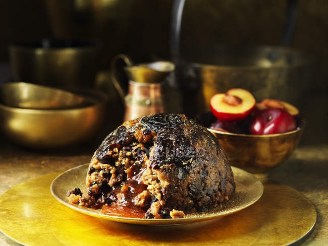 Sugar plum pudding on golden plate, fresh plums in background — Stock Photo