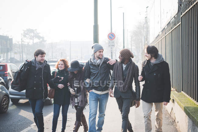 Seis amigos adultos jóvenes charlando mientras pasean por la calle de la ciudad - foto de stock