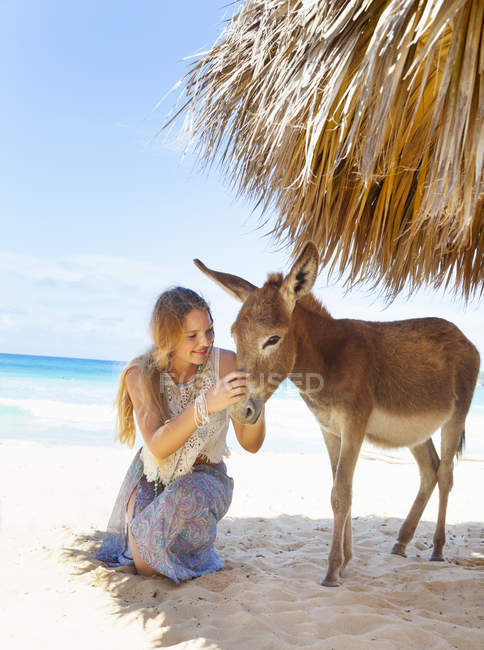 Mujer joven arrodillada para acariciar burro en la playa, República Dominicana, El Caribe - foto de stock