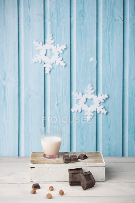 Chocolate cubes, hazelnuts, glass of milk, paper snowflakes on blue wall — Stock Photo
