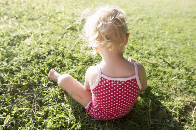 Rear view of female toddler wearing red spotted bathing costume sitting on grass — Stock Photo