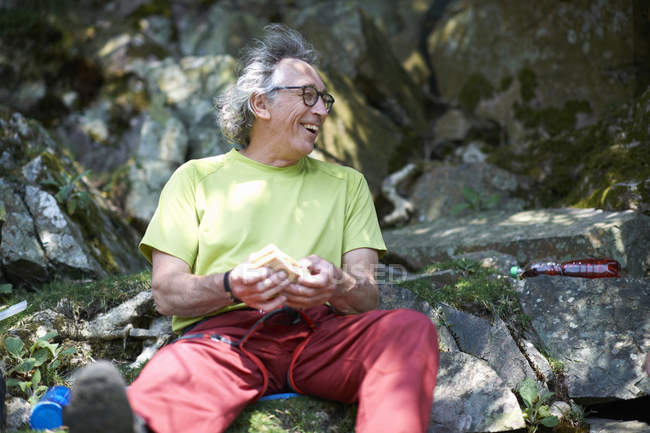 Hiker sitting on rocks holding sandwich looking away smiling — Stock Photo