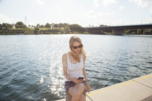 Jeune femme assise au bord de la rivière lisant des textes sur smartphone, île du Danube, Vienne, Autriche — Photo de stock
