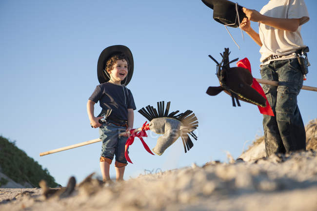 Dois irmãos vestidos de cowboys com cavalos de passatempo na areia — Fotografia de Stock