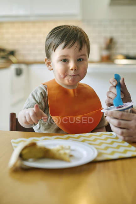Niño sentado en la mesa siendo alimentado con yogur por la madre - foto de stock