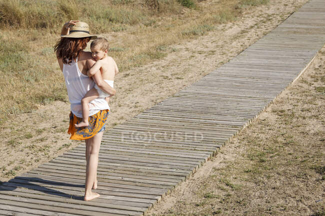 Rear view of mother walking down boardwalk carrying son in arms — Stock Photo