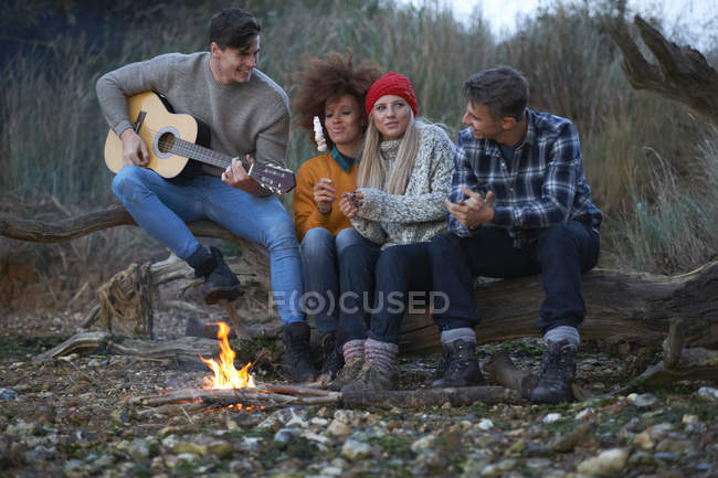 Vier junge erwachsene Freunde spielen in der Abenddämmerung am Lagerfeuer am Strand Gitarre — Stockfoto