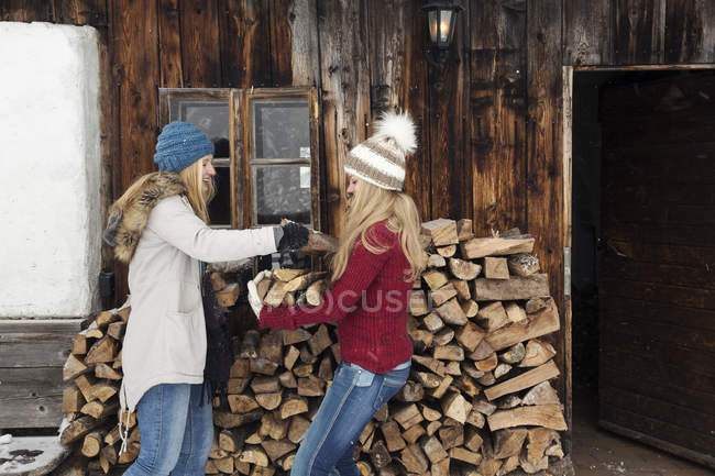 Dos amigas jóvenes apilando troncos fuera de la cabaña - foto de stock