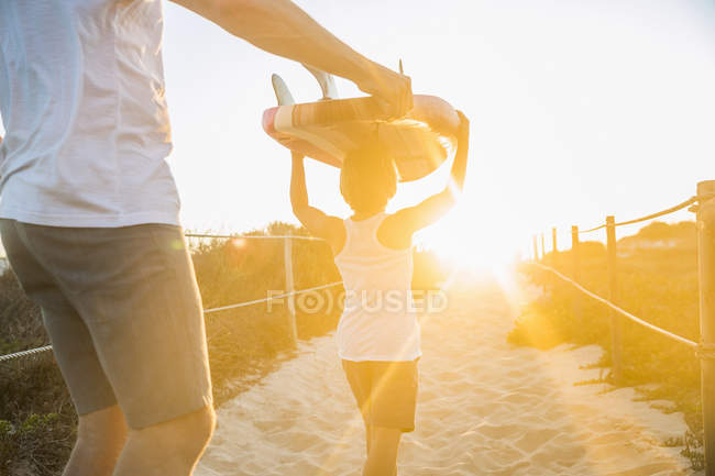Вид сзади на отца и сына на пляже, держащихся за руки, несущих доску для серфинга над головой — стоковое фото