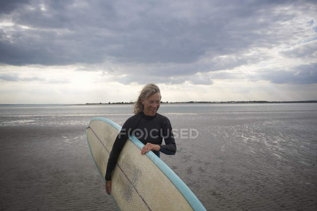 Старша жінка, що йде з моря, носить дошку для серфінгу — стокове фото