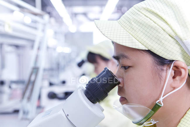 Jovem mulher usando microscópio na estação de verificação de qualidade na fábrica produzindo placas de circuito eletrônico flexível. Planta está localizada no sul da China, em Zhuhai, província de Guangdong — Fotografia de Stock