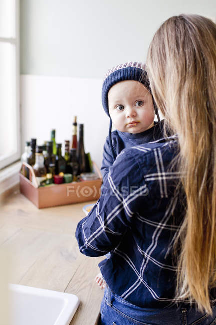 Femme portant un bébé fils portant un chapeau en tricot dans la cuisine — Photo de stock