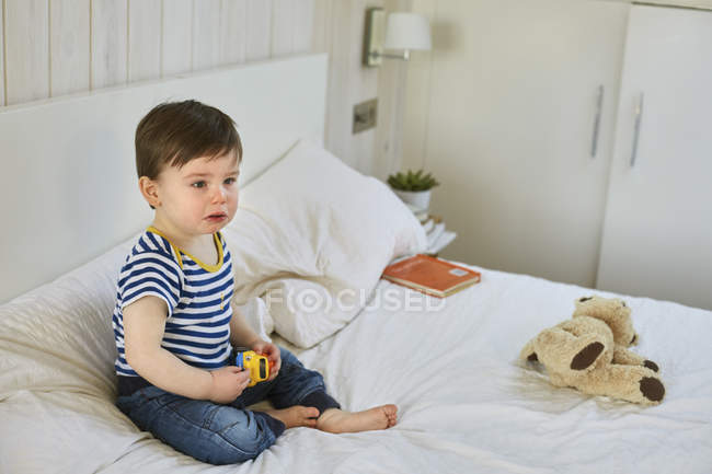 Triste bébé garçon assis sur lit tenant voiture jouet, regardant loin — Photo de stock