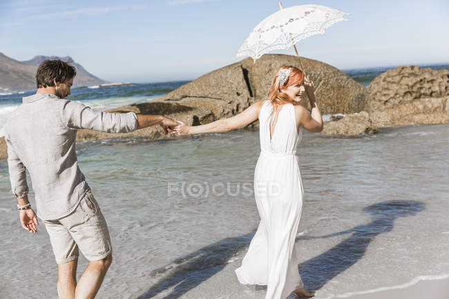 Couple tenant la main, marchant sur le littoral tenant parapluie regardant par-dessus l'épaule souriant — Photo de stock