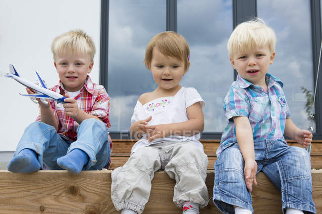 Niño con avión de juguete y dos niños pequeños sentados en el patio - foto de stock
