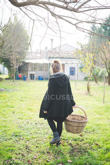 Full length rear view of young woman in garden carrying wickerwork basket — Stock Photo