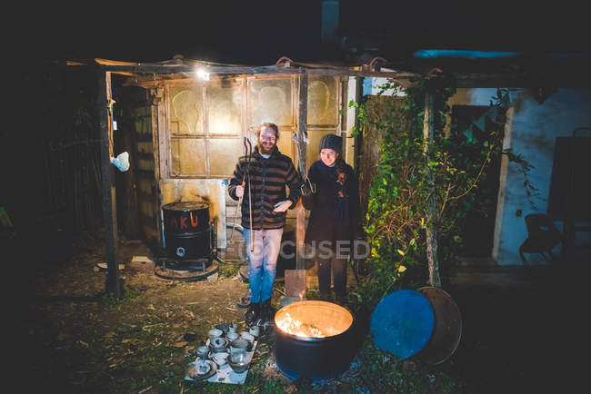 Casal na frente de galpão segurando pinças, com cerâmica pelo fogo no barril — Fotografia de Stock