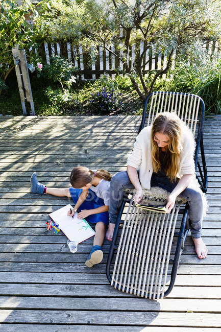 Mère utilisant une tablette, fille dessinant sur une terrasse en bois — Photo de stock