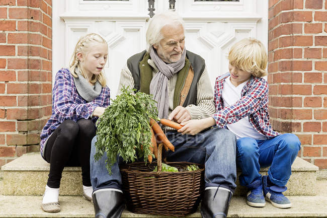 Grandfather sitting with grandchildren on doorstep with carrots — Stock Photo