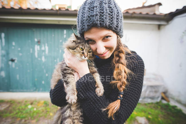 Giovane donna che indossa cappello a maglia coccole gatto, guardando la fotocamera sorridente — Foto stock