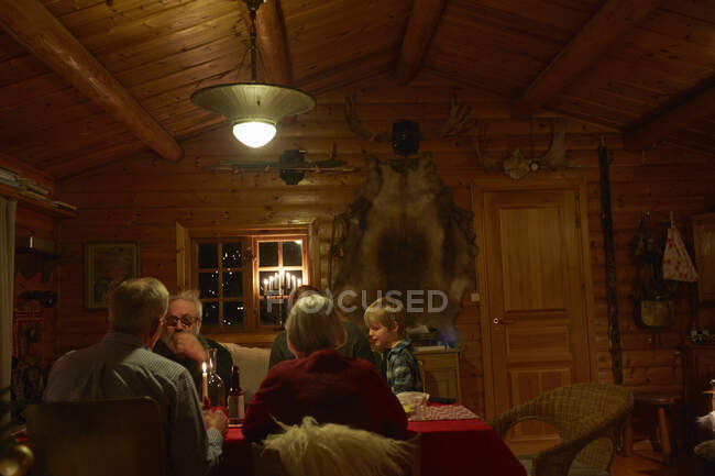 Familia de tres generaciones sentada hablando en la mesa de Navidad en la cabaña de madera por la noche - foto de stock