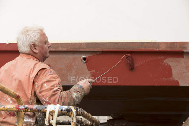 Senior male ship painter roller painting ship hull red in ship painters yard — Stock Photo
