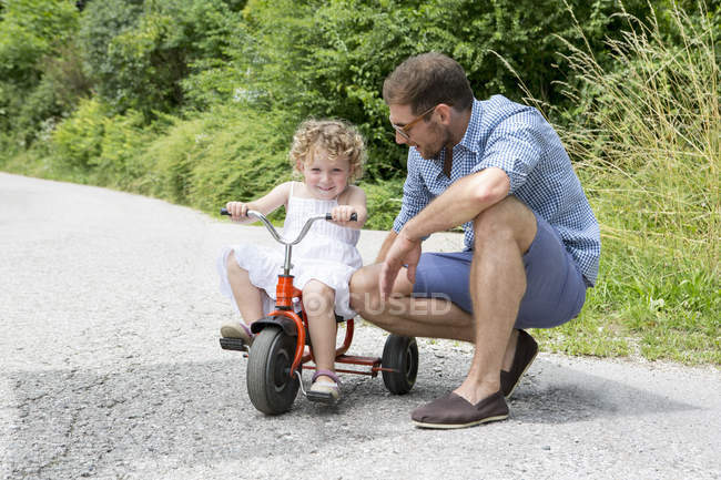 Girl riding tricycle with father on rural road — Stock Photo