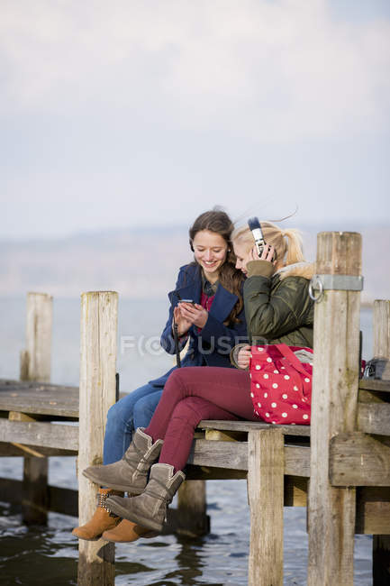 Teenage girls sitting on jetty and listening music — Stock Photo