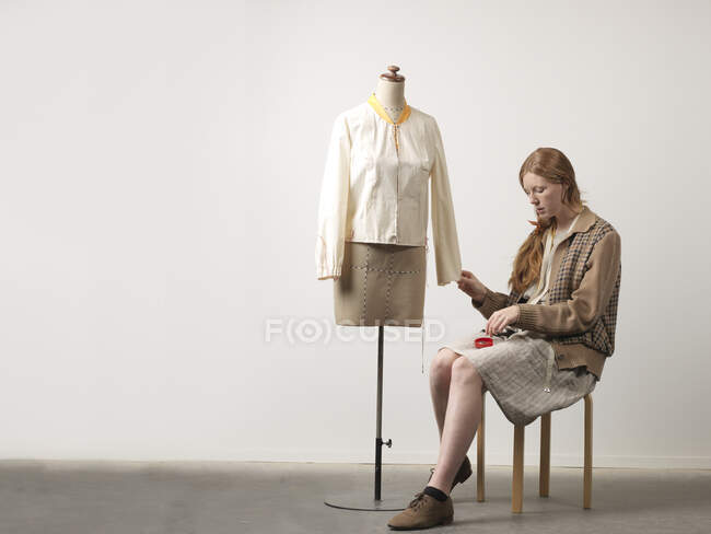 Joven diseñadora de moda sentada en el taburete sujetando la blusa en el maniquí de la modista - foto de stock