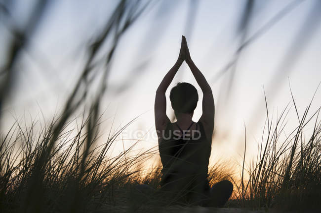 Rear view of mid adult woman practicing yoga in silhouetted  long grasses at sunset — Stock Photo