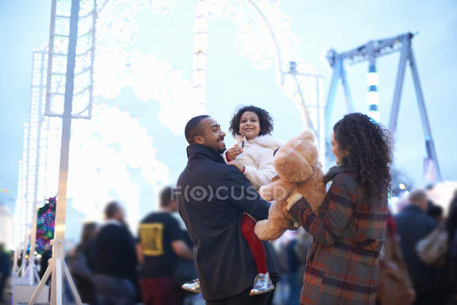 Madre y padre en el parque de atracciones llevando a una chica sonriente y un oso de peluche - foto de stock
