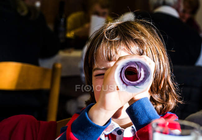 Retrato de niño mirando a través de papel enrollado en la cafetería - foto de stock