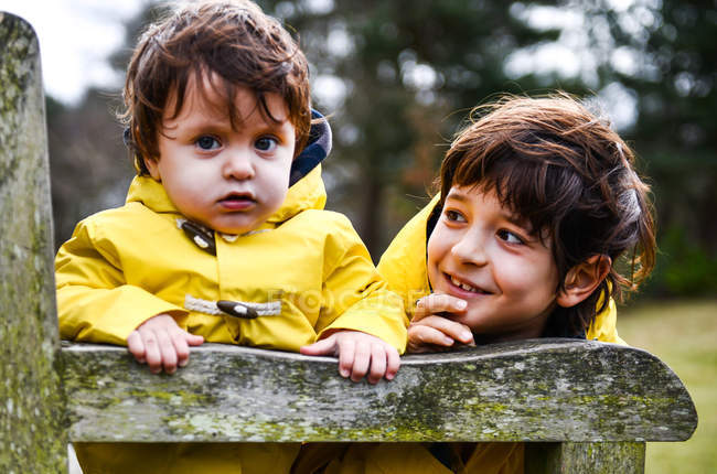 Retrato de bebé y hermano mayor en anoraks amarillos en el banco del parque - foto de stock