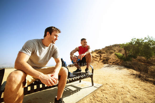 Zwei männliche Freunde, die auf einer Bank auf einer Klippe sitzen und die Aussicht betrachten — Stockfoto