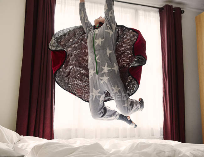 Menino vestindo capa pulando na cama — Fotografia de Stock