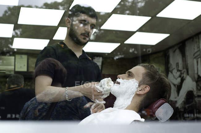 Vista através da janela do jovem na barbearia aplicando creme de barbear no rosto dos clientes — Fotografia de Stock