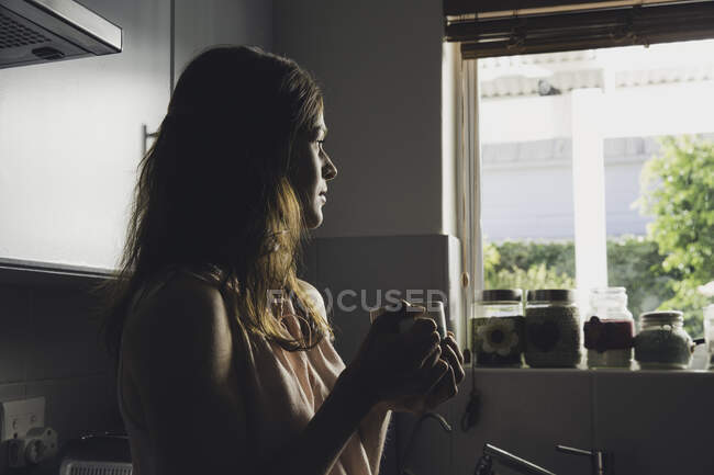Mujer joven tomando un café mirando a través de la ventana de la cocina - foto de stock