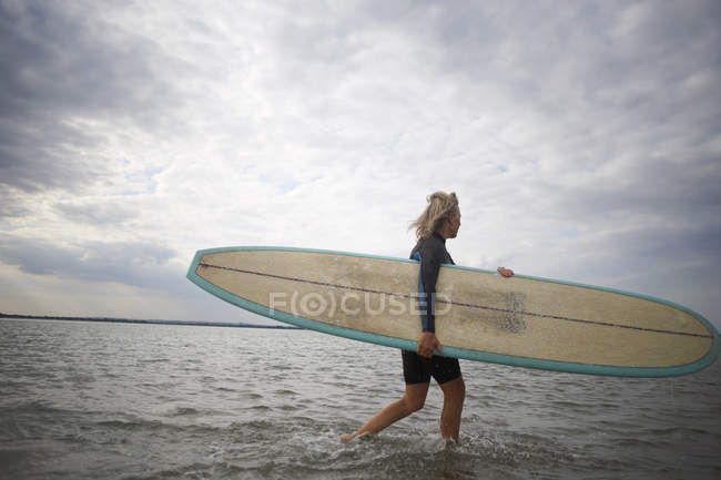 Старша жінка, що йде з моря, носить дошку для серфінгу — стокове фото