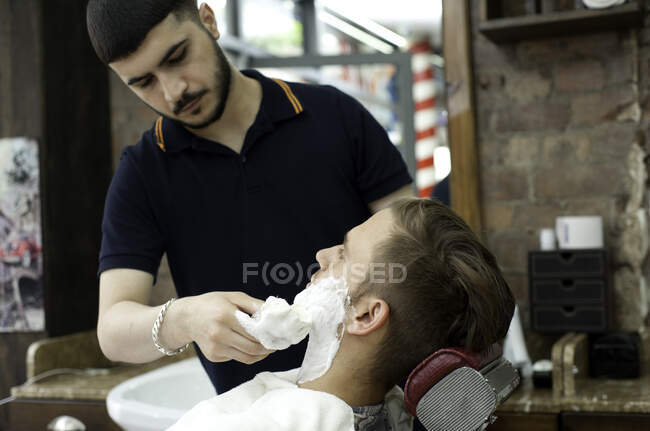 Jeune homme dans le salon de coiffure appliquant la crème à raser aux clients visage — Photo de stock