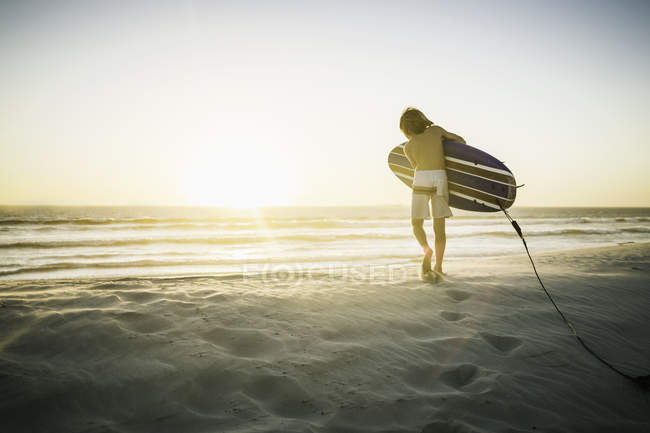 Jeune garçon sur la plage, portant une planche de surf, marchant vers la mer, vue arrière — Photo de stock