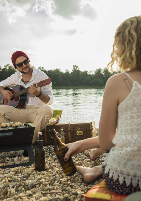 Jeune homme assis au bord du lac jouant de la guitare — Photo de stock