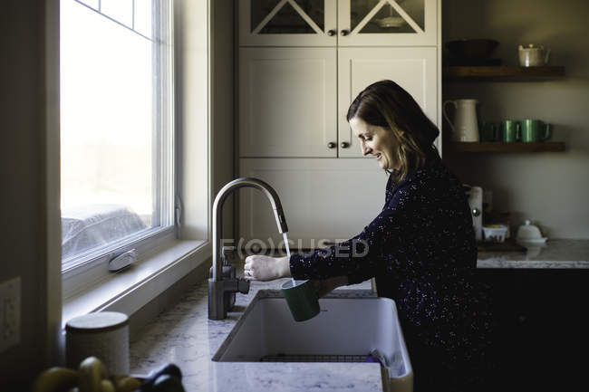 Mujer llenando taza en fregadero de cocina - foto de stock