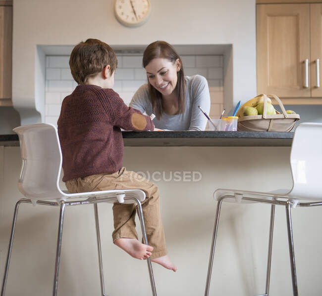 Menino sentado no banquinho com a mãe na cozinha e desenho — Fotografia de Stock