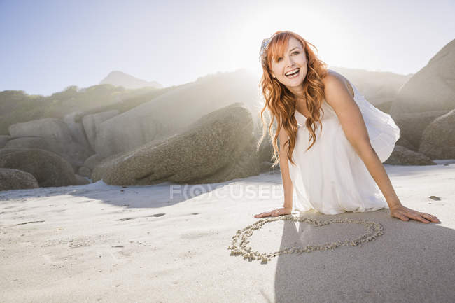 Жінка присіла на пляжі, малюючи форму серця в піску, дивлячись геть посміхаючись — стокове фото