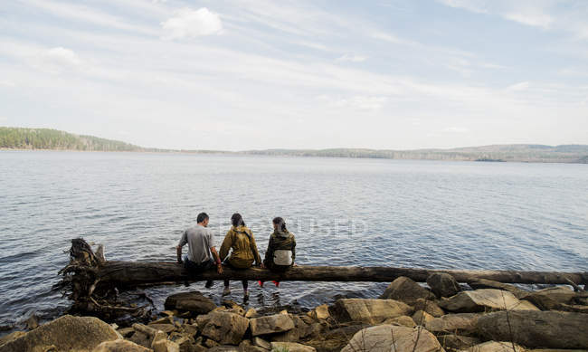 Three hiking friends sitting in a row on fallen tree at lakeside — Stock Photo