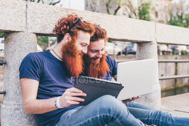 Young male hipster twins with red hair and beards sitting on bridge browsing digital tablet — Stock Photo