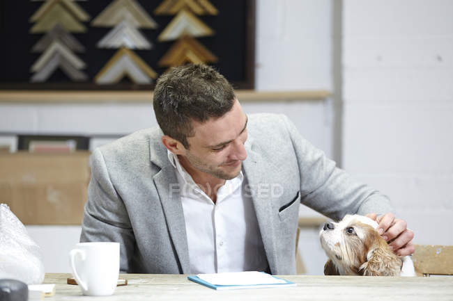 Mittlerer erwachsener Mann streichelt Hund am Schreibtisch in Bilderrahmenwerkstatt — Stockfoto