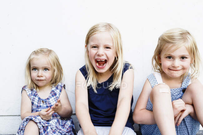 Retrato de tres hermanas jóvenes sentadas frente a la pared blanca - foto de stock