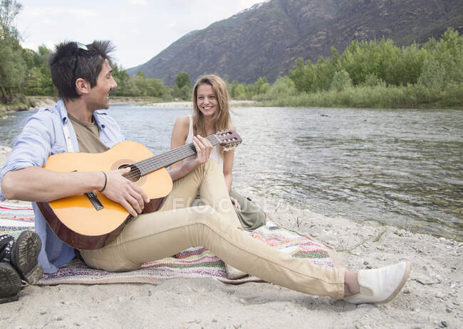 Друзі сидять біля річки, грають на гітарі — стокове фото