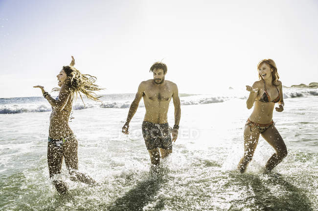 Three adult friends wearing bikinis and swimming shorts splashing in sea, Cape Town, South Africa — Stock Photo